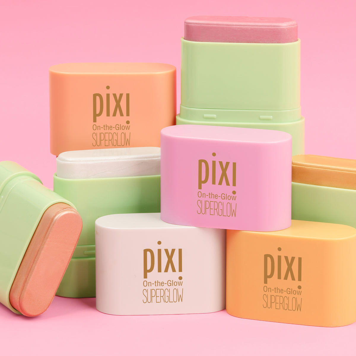 A group of Pixi On-the-Glow SuperGlow Petite stick products in pastel shades of pink, orange, green, and white, arranged on a solid pink background.