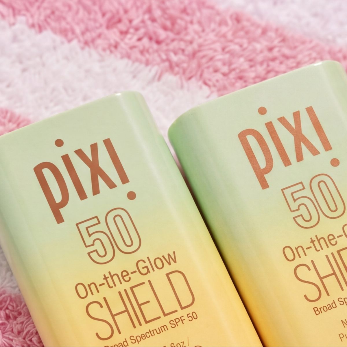 Two Pixi On-the-Glow Shield Broad Spectrum SPF 50 sunscreen bottles with a light green to pale yellow gradient, resting on a pink textured towel.