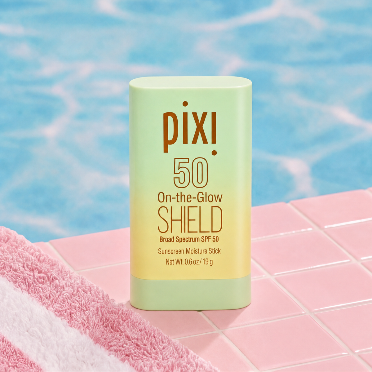 Pixi On-the-Glow Shield SPF 50 sunscreen stick standing on pink tiled decking with a blue swimming pool in the background and a pink towel in the foreground.