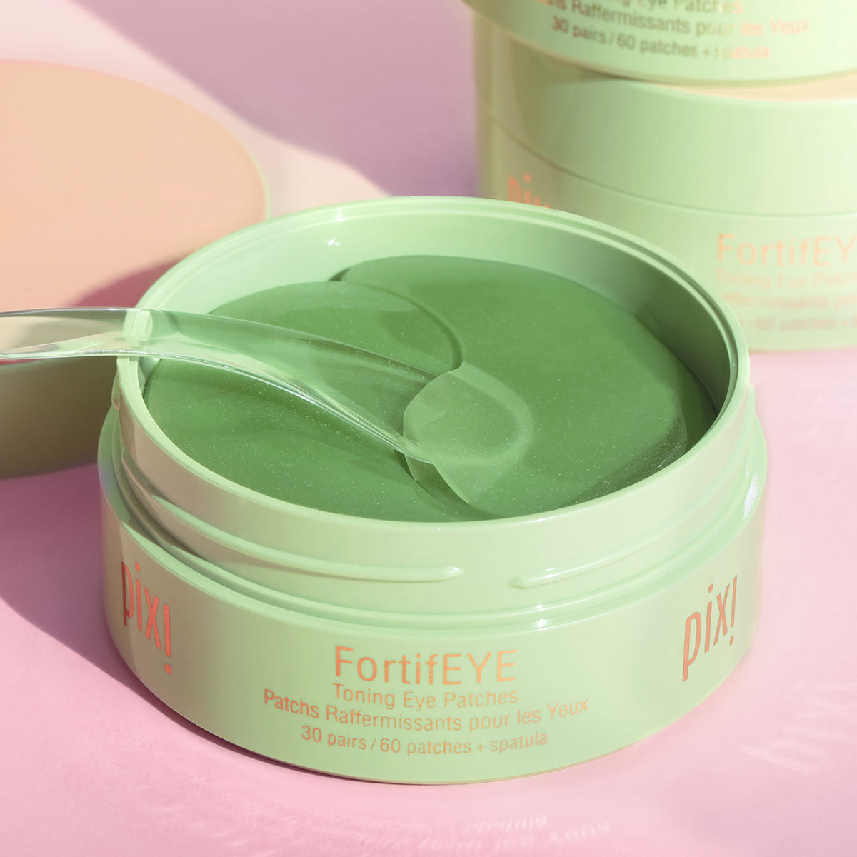 An open round jar of Pixi FortifEYE toning eye patches in a mint green shade, with a clear plastic spatula resting inside. The background is a soft pink with a second closed jar blurred in the background.